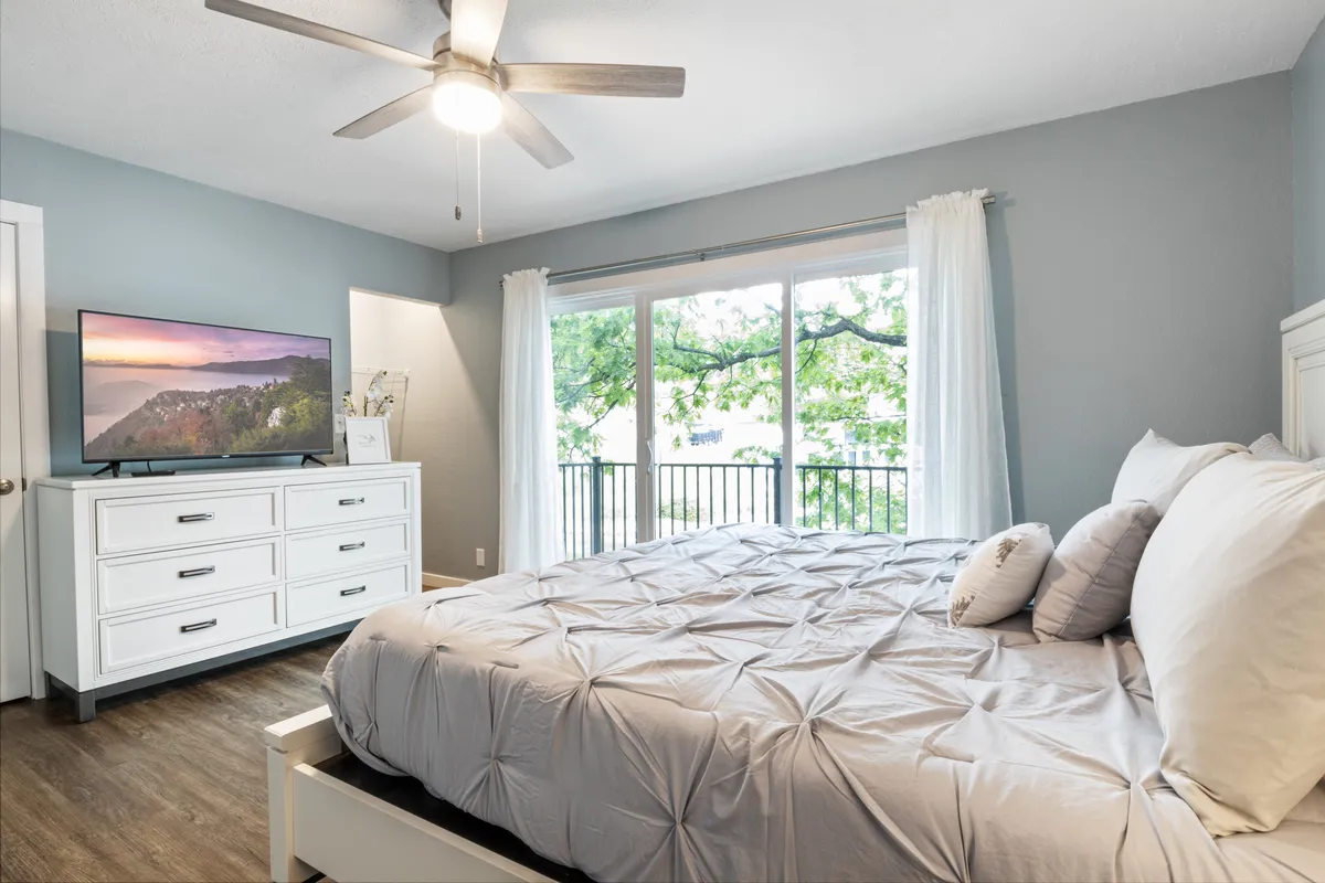 A tranquil bedroom filled with natural light from a large window, featuring a comfortable bed with decorative pillows and a television that offers a scenic view. The space is styled for relaxation, encouraging restful sleep and moments of quiet retreat.