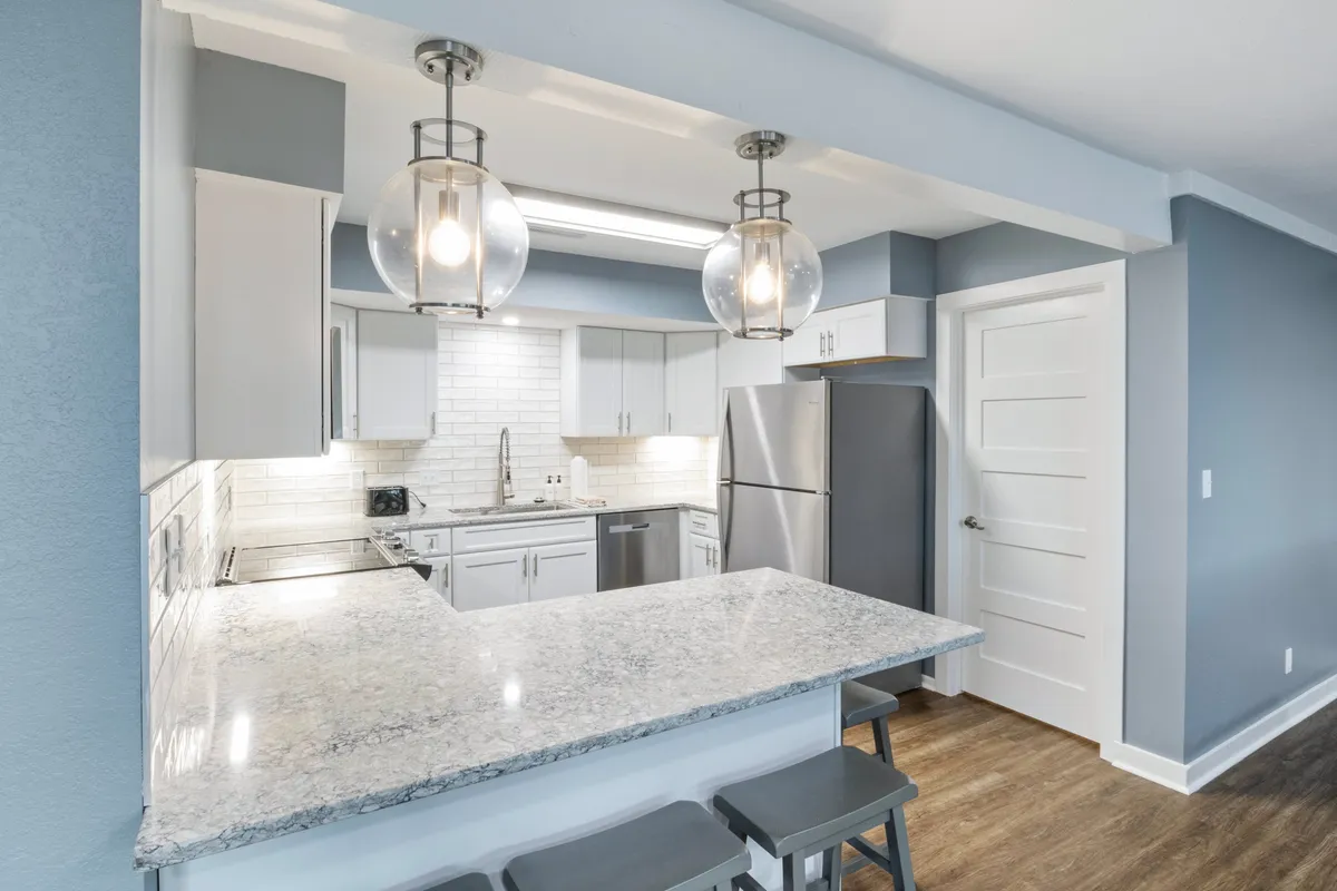 The kitchen in this image is bright and inviting, featuring a granite countertop, pendant lighting, stainless steel appliances, and white cabinetry. The space is clearly designed for both cooking and casual dining, with plenty of room for family or friends to gather.