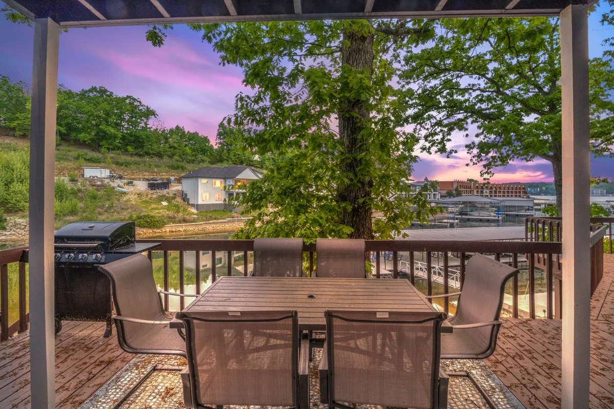 This exterior image captures a dining table with reclining chairs and a grill, all set on a deck overlooking the lake. The distant water view and a colorful sunset enhance the atmosphere, while the setup suggests a perfect spot for alfresco dining and relaxation.