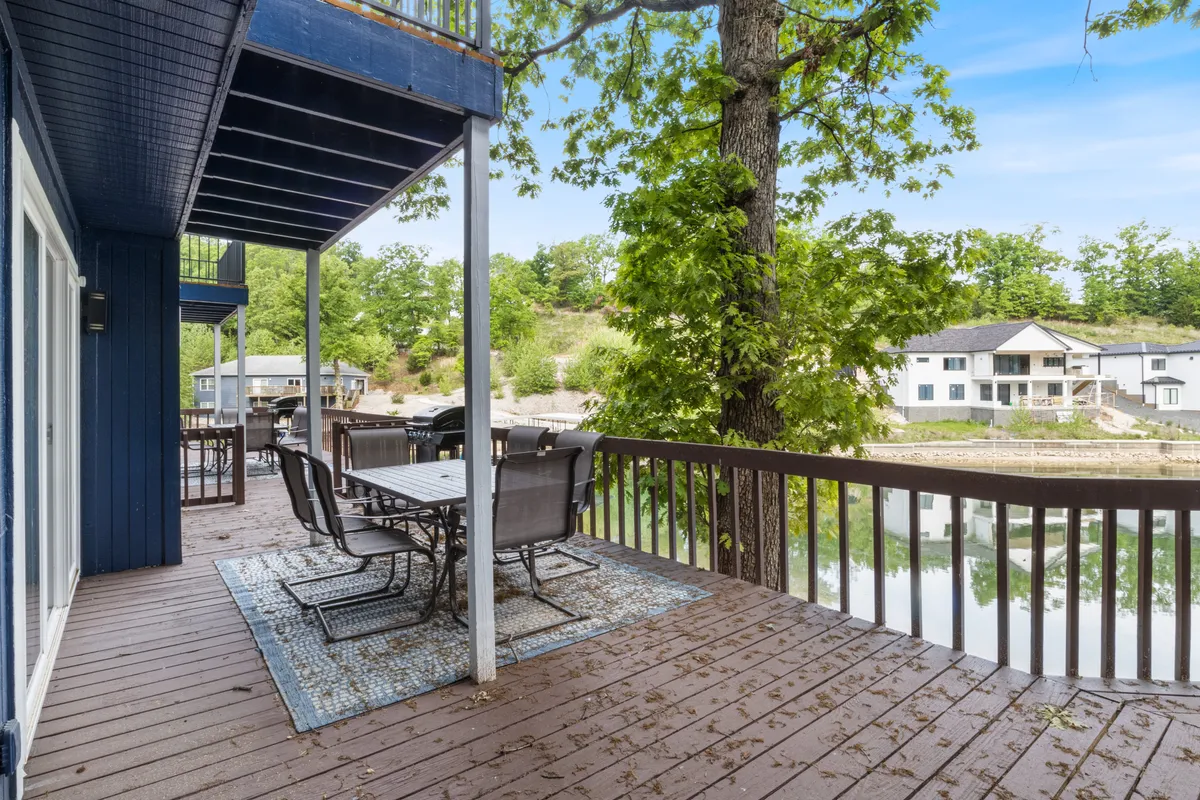An inviting outdoor dining area overlooks the lake, surrounded by trees and shaded by the natural landscape. The setup includes a table, comfortable outdoor seating, and views of the shimmering water, ideal for gatherings and unwinding by the water's edge.