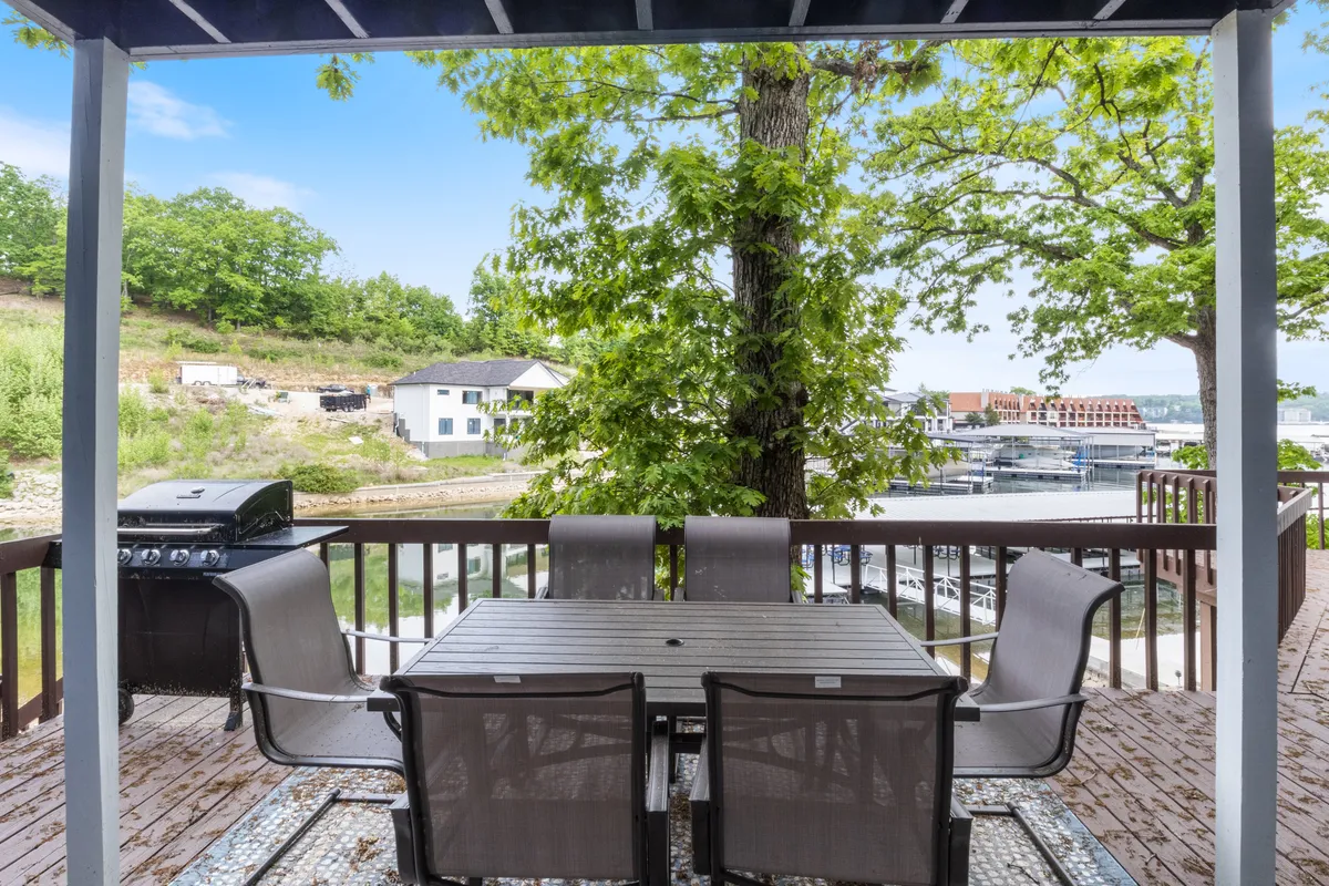 A deck with a dining table and grill offers panoramic views of the marina, with boats visible in the background and lush greenery surrounding the space. The setup is perfect for enjoying meals outdoors while watching lake activity.