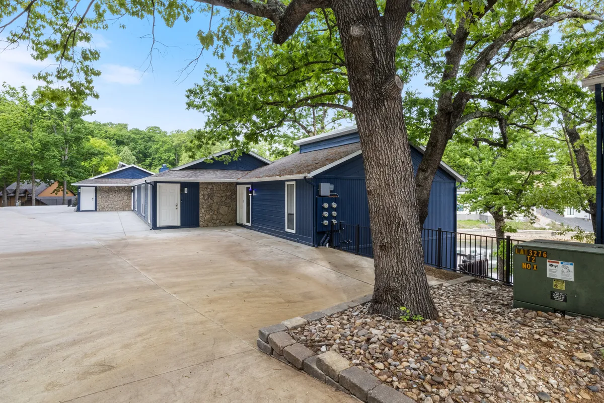 A welcoming exterior view of the Cove Retreat, featuring a charming blue house with stone accents set amidst mature trees and a spacious driveway. The large tree offers shaded spots, and the front area suggests plenty of room for outdoor activities and gatherings.