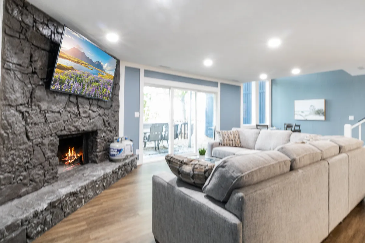 This image features a cozy, modern living area with a stone fireplace, a large TV set up for movie nights, a plush sectional sofa perfect for group lounging, and sliding glass doors that flood the space with natural light and offer easy access to the outdoors.