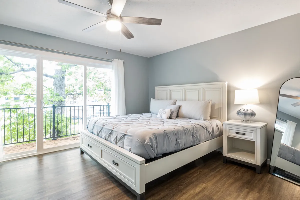 A cozy bedroom with a plush bed, nightstand, lamp, mirror, and balcony access. The view from the balcony offers a peaceful start to the day, while soft lighting and thoughtful details create an inviting atmosphere perfect for unwinding.