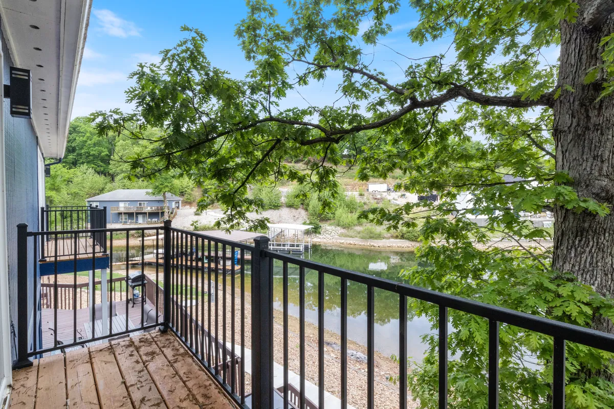 A peaceful lakeside view seen from a balcony, framed by a railing and tree branches. The calm water and distant houses evoke a tranquil setting perfect for family fun and safe swimming. The image gives a sense of the serene, wave-free cove mentioned in the section.