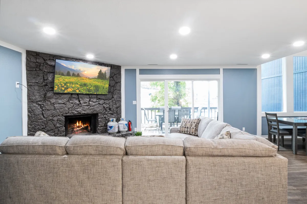 The living area is shown with a large sectional sofa, a welcoming fireplace, a TV displaying a beautiful nature scene, and glass doors that open to a visible dining area and deck, allowing for effortless flow between indoor and outdoor spaces.