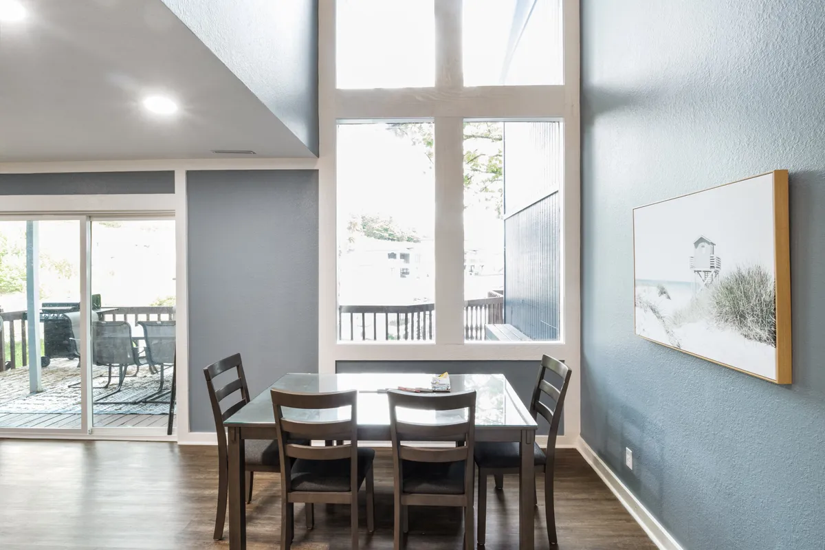 This image focuses on a dining table set beside large windows, with artwork on the wall and soft natural light streaming in. The setup suggests a space where family meals and casual gatherings are enhanced by sunlight and views.